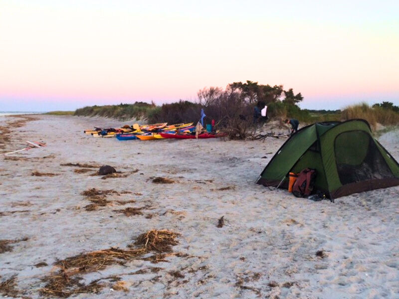 The image shows a campsite on a sandy beach. A green tent is pitched in the foreground, with a backpack next to it. In the background, there's a collection of colorful kayaks and some vegetation. The sky is a soft gradient of colors, suggesting it's either sunrise or sunset. The scene conveys a sense of outdoor adventure and tranquility.
